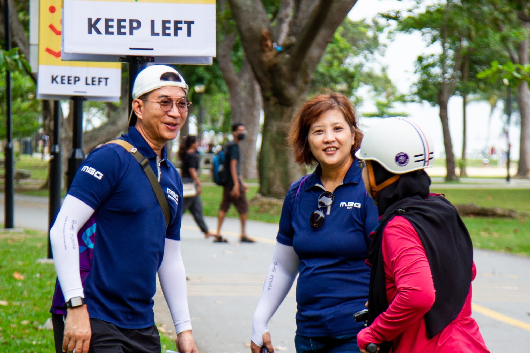 Two people in blue shirts chat with a helmeted person in pink beside a park path and KEEP LEFT signs.